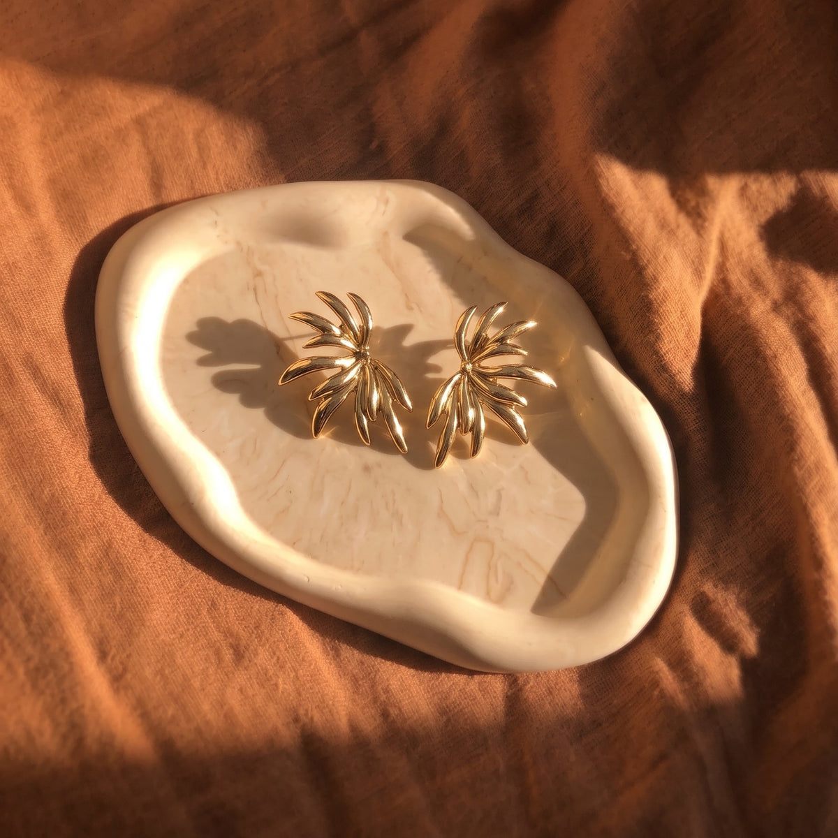 A pair of gold-toned, palm-leaf-shaped stud earrings displayed on an organic-shaped cream ceramic tray, set against a draped rust-orange linen fabric background.