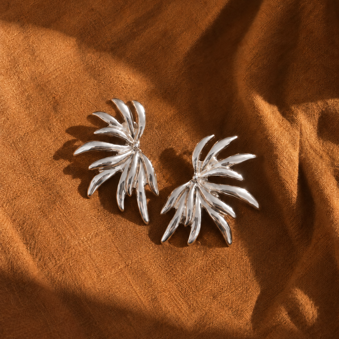 A pair of silver-toned palm leaf stud earrings resting on draped brown fabric.