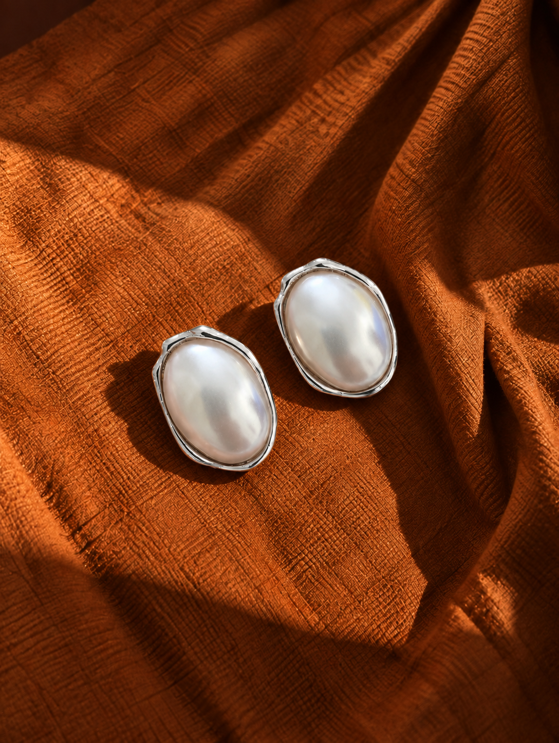 Two oval pearl stud earrings with silver settings, resting on a draped rust-orange textured fabric.