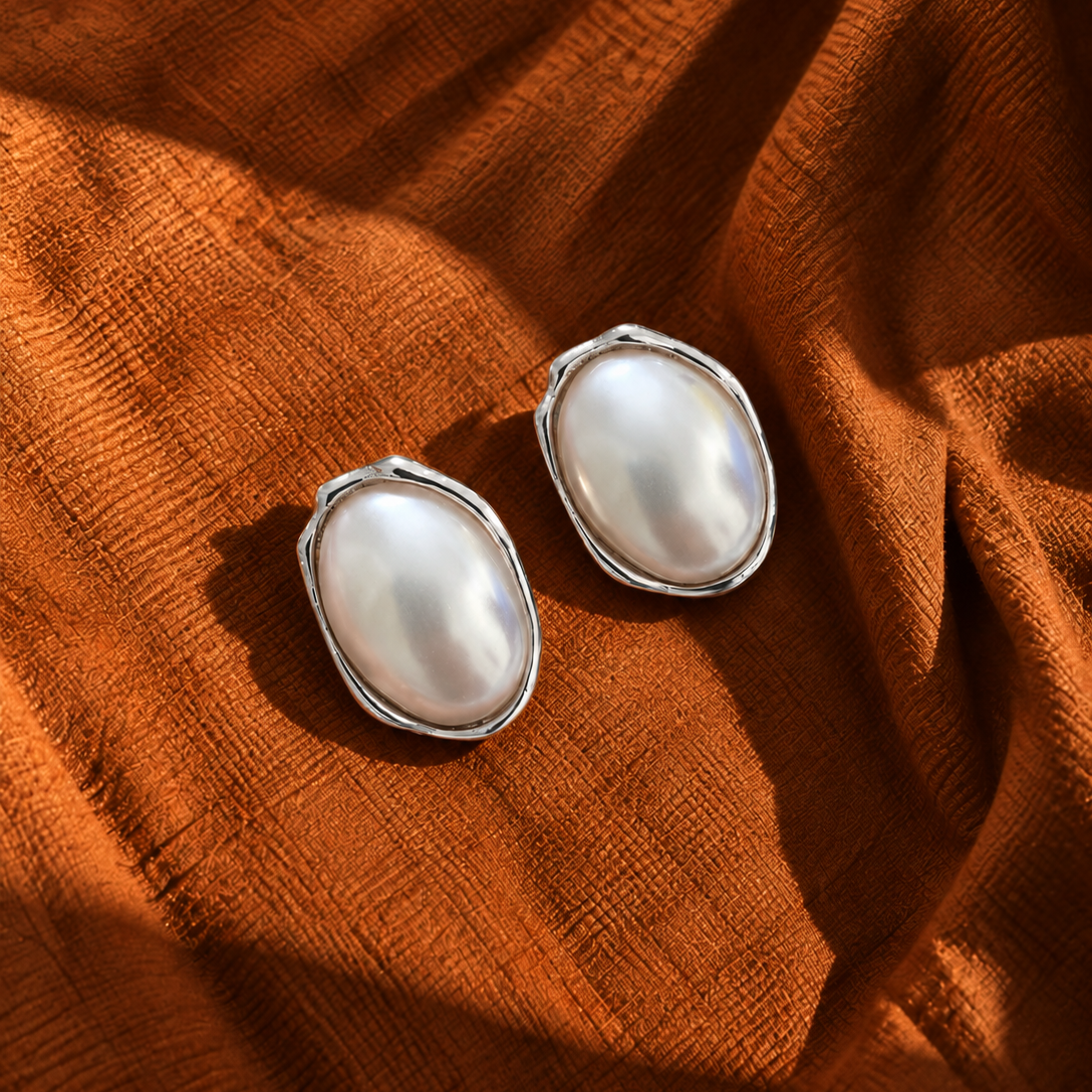 Two oval pearl stud earrings with silver settings, resting on a draped rust-orange textured fabric.