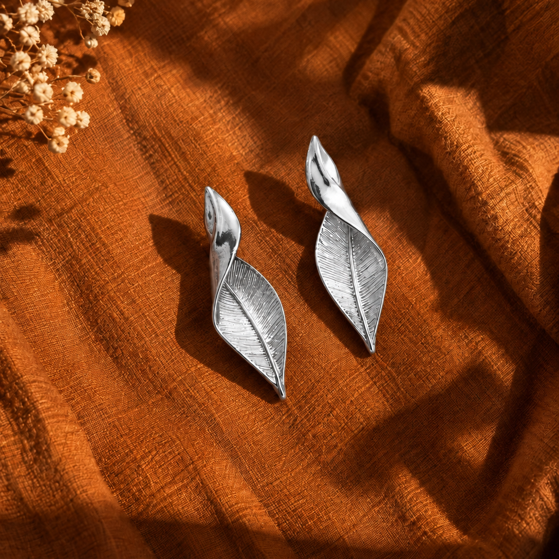 A pair of silver leaf-shaped drop earrings displayed on a textured rust-orange fabric background with dried flowers in the top-left corner.