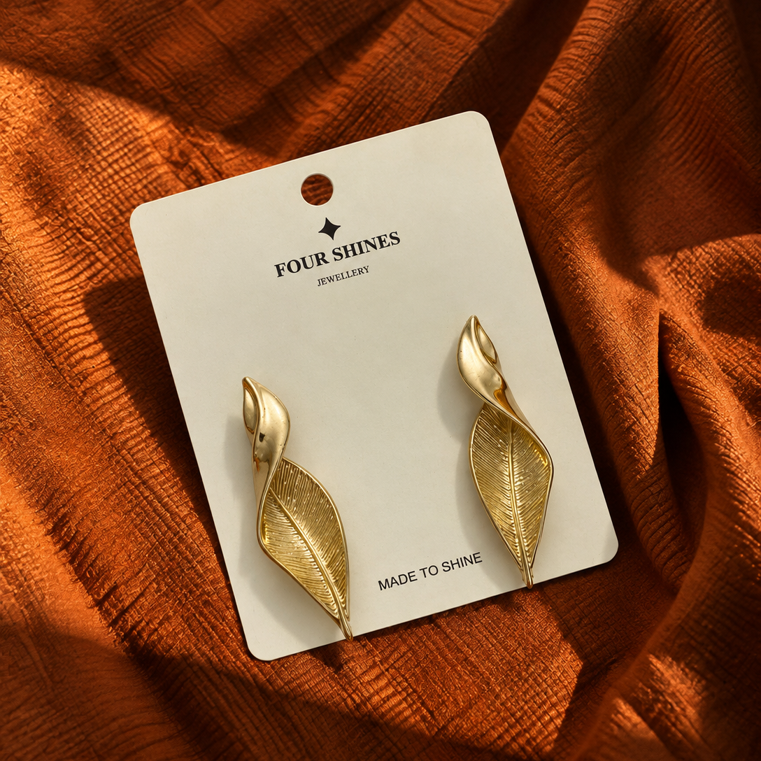 A pair of gold leaf-shaped dangle earrings on a white display card, placed on a textured rust-orange fabric background. The card is branded 'FOUR SHINES JEWELLERY'.