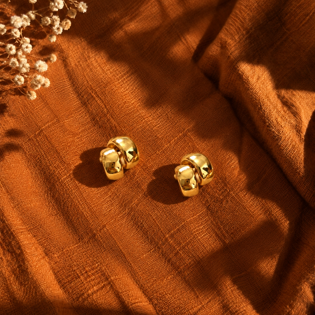 A pair of gold-toned double-huggie hoop earrings resting on a textured rust-orange fabric, styled with dried white flowers in the top-left corner under warm, dramatic sunlight.