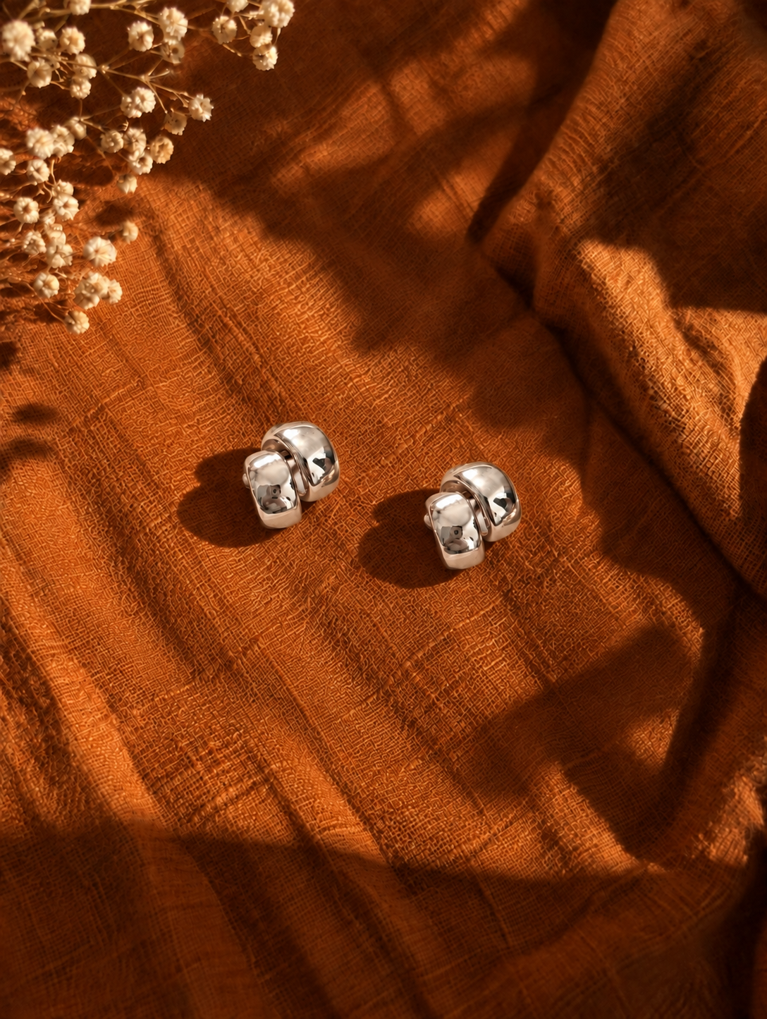 A pair of polished silver stud earrings with a textured, rounded cube design, resting on a draped rust-orange linen fabric with dried flowers in the corner.