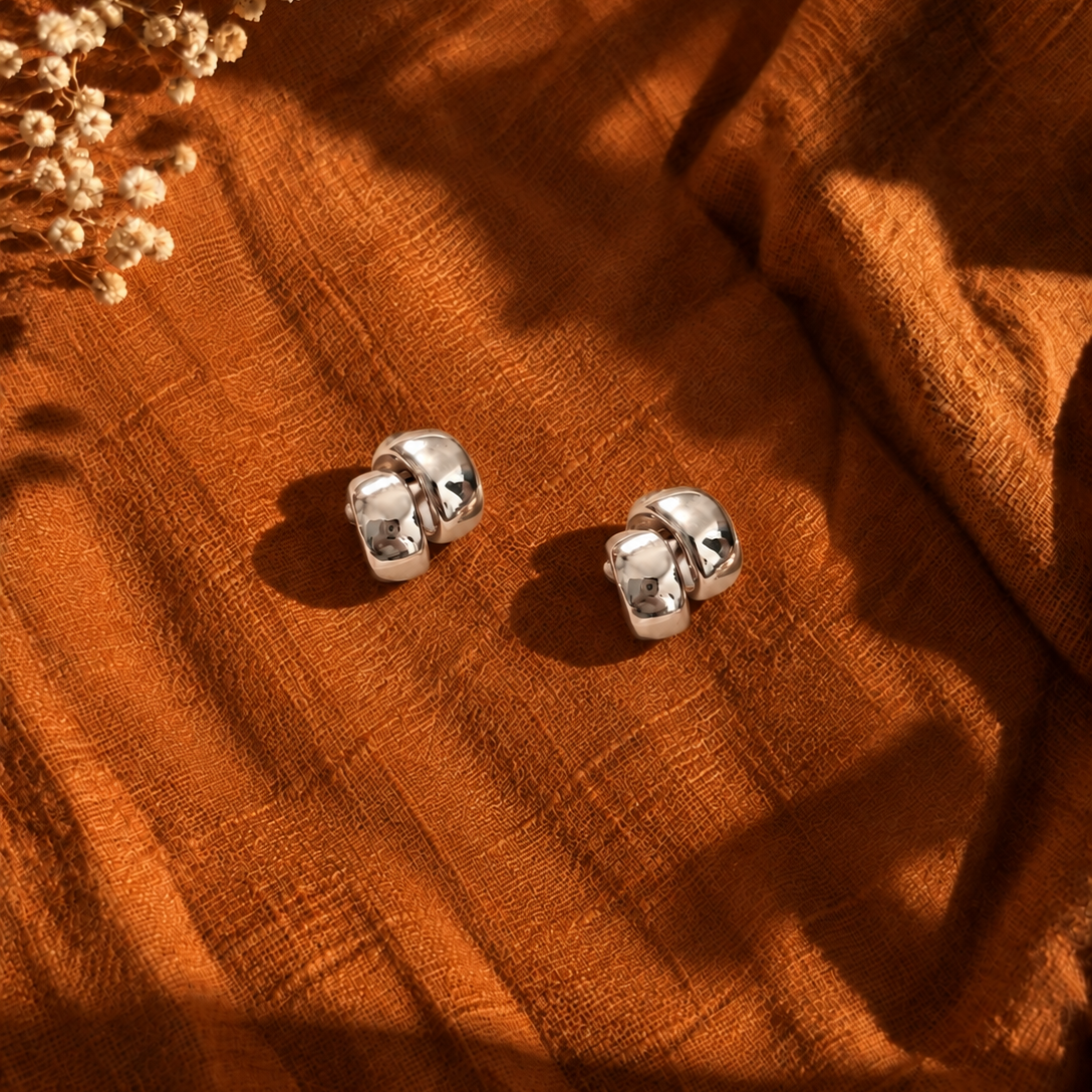 A pair of polished silver stud earrings with a textured, rounded cube design, resting on a draped rust-orange linen fabric with dried flowers in the corner.