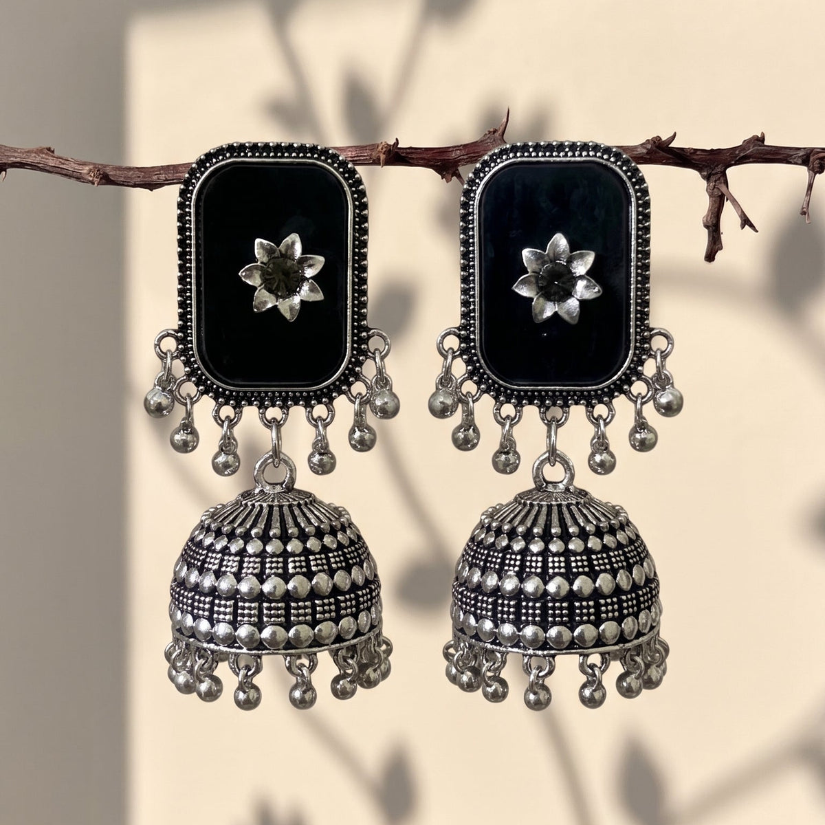 A pair of black and silver jhumka earrings hanging from a thin branch. Each earring features a rectangular black face with a silver floral motif, adorned with dangling silver beads and bell-shaped jhumka drops with intricate silver patterns.