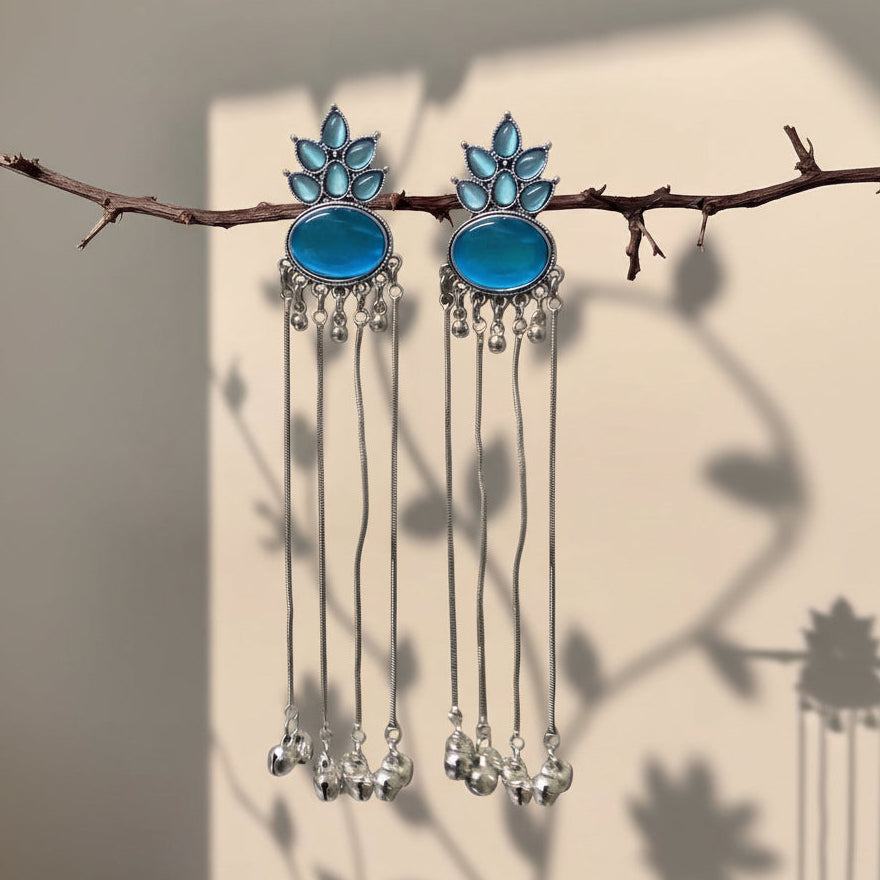 A pair of silver-toned dangle earrings with oval blue stones and leaf-shaped blue accents, hanging from a thin branch against a beige wall with soft leaf shadows.