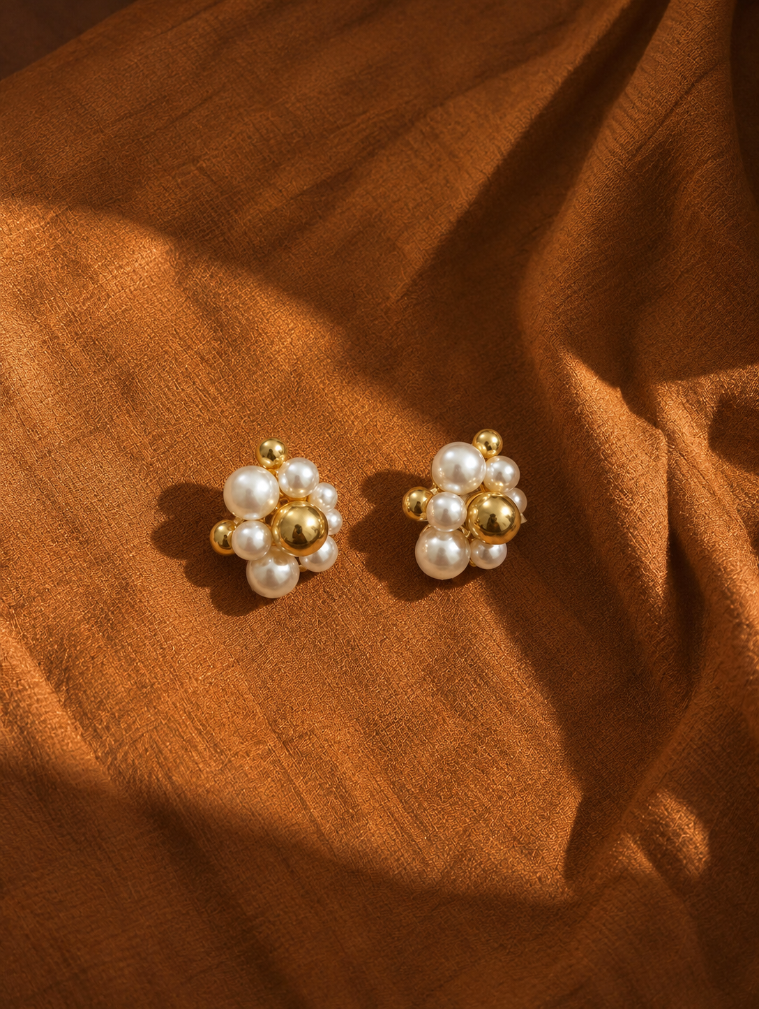A pair of gold and pearl stud earrings arranged on draped brown fabric.