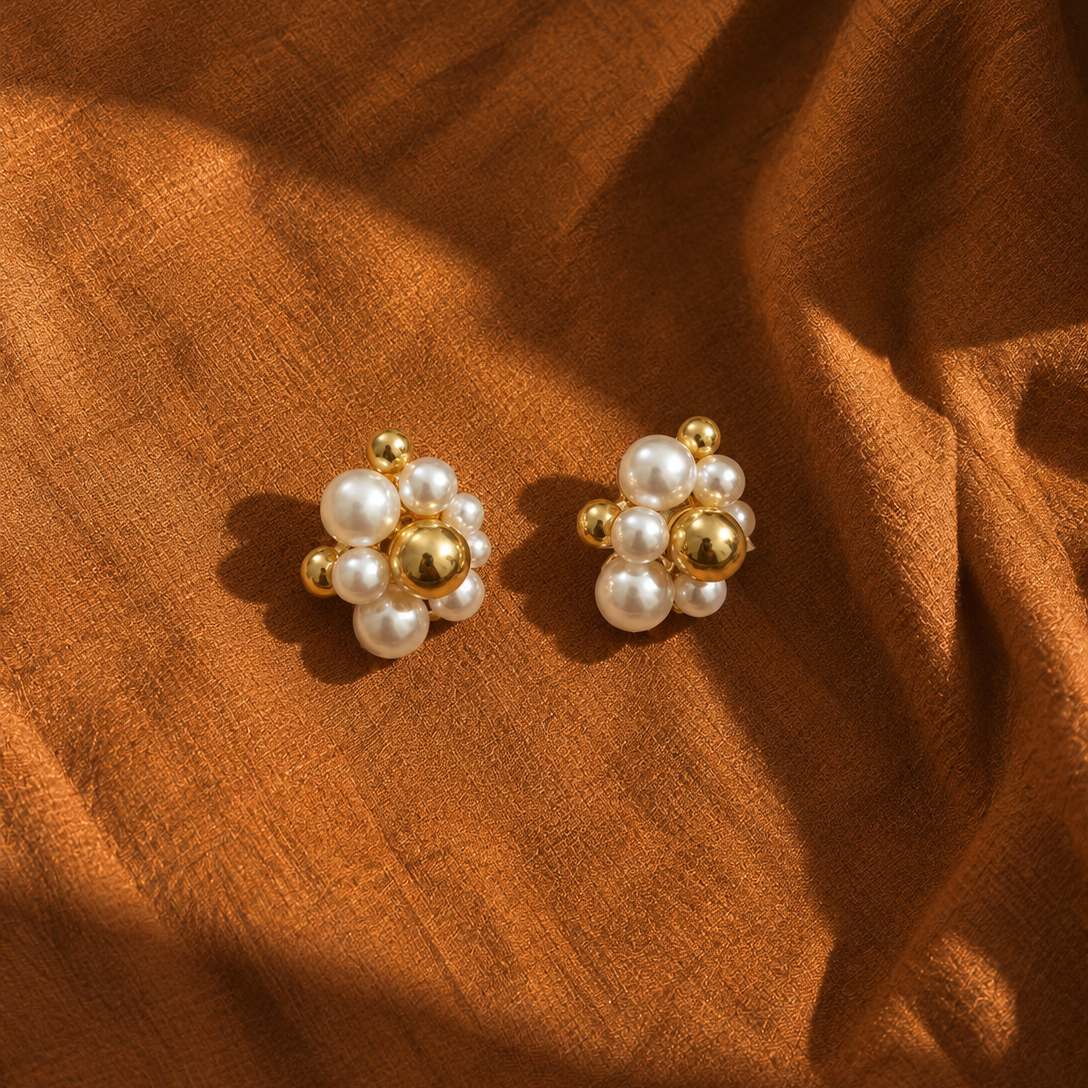 A pair of gold and pearl stud earrings arranged on draped brown fabric.