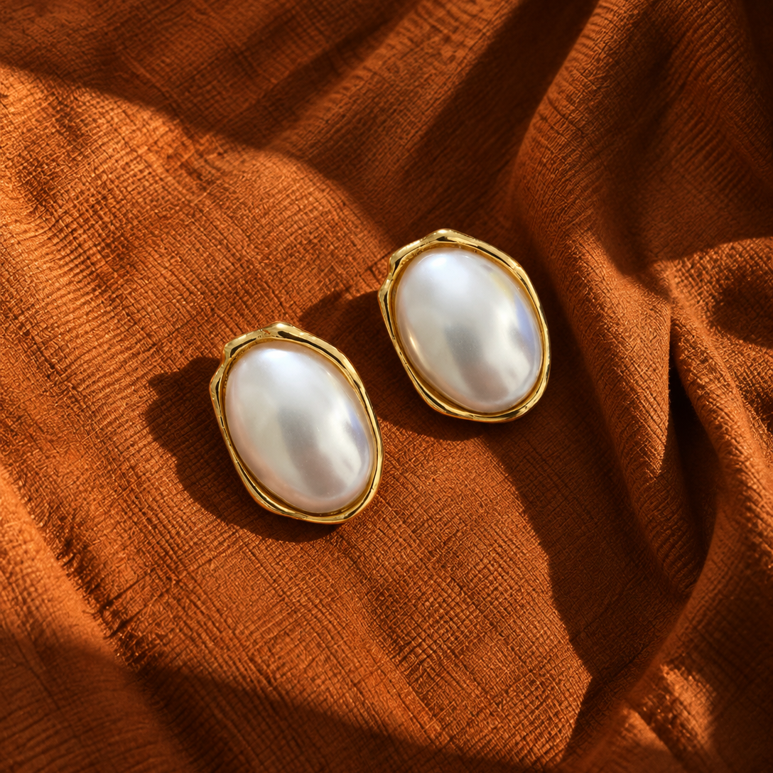 A pair of gold-toned oval earrings with large white pearl-like centers, displayed on draped rust-orange fabric.