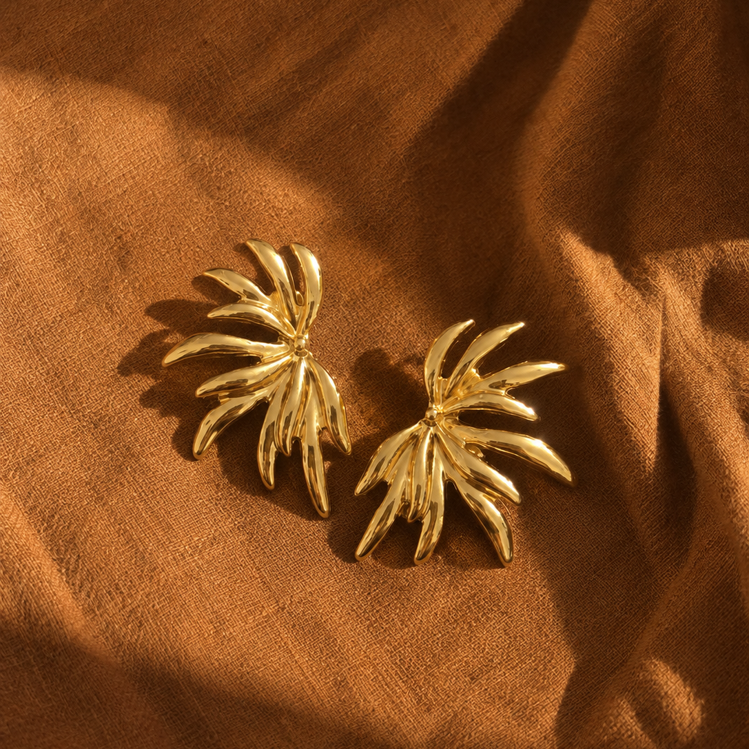 A pair of gold-toned, palm-leaf-shaped earrings resting on draped brown fabric under warm, directional sunlight.