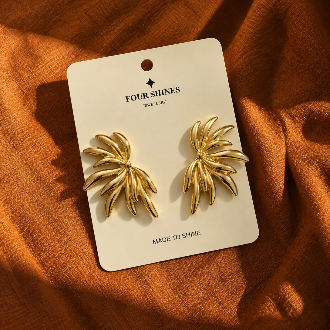A pair of gold leaf-shaped stud earrings on a white display card labeled 'FOUR SHINES JEWELLERY', placed on a textured rust-orange fabric background.