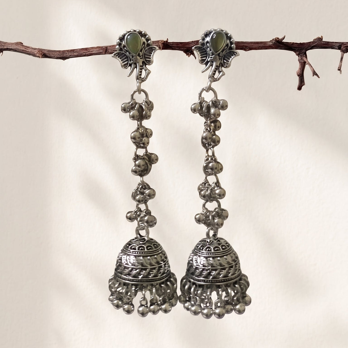 A pair of traditional silver-toned jhumka earrings with green stone accents, hanging from a thin brown branch against a warm off-white background.