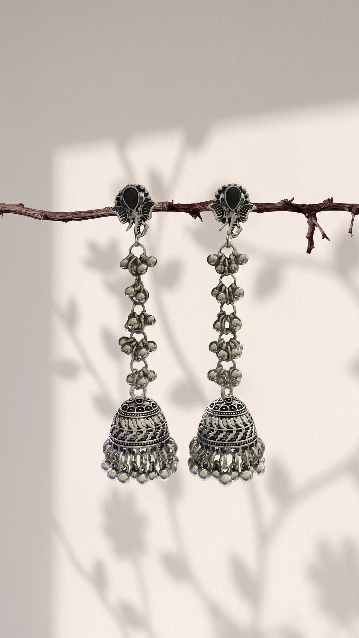 A pair of silver-toned Indian jhumka earrings hanging from a thin brown branch against a warm off-white background with soft leaf shadows.