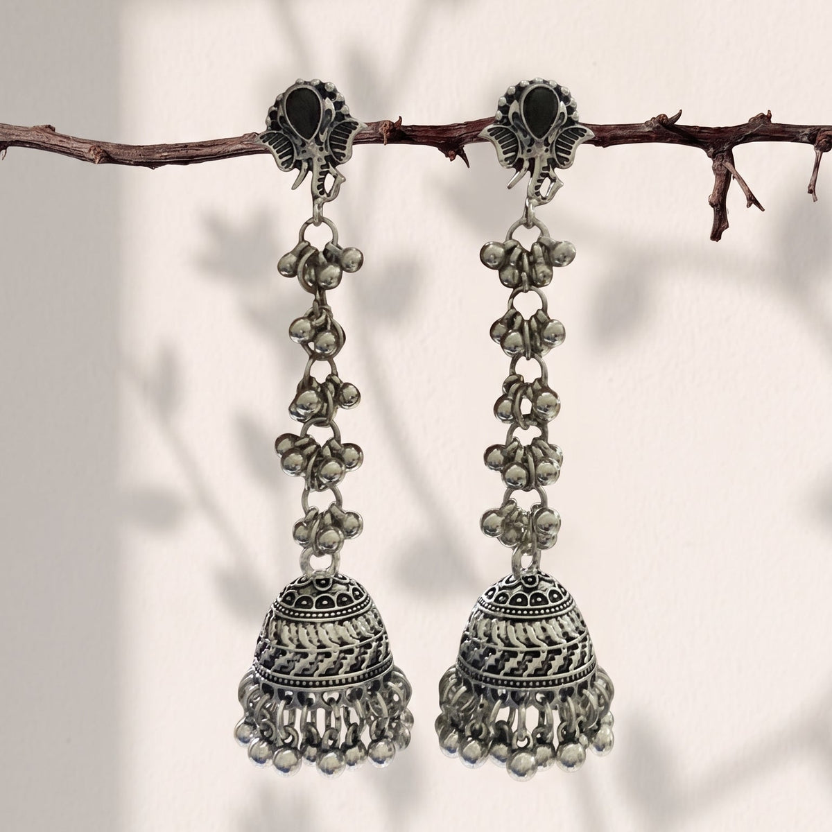 A pair of silver-toned Indian jhumka earrings hanging from a thin brown branch against a warm off-white background with soft leaf shadows.