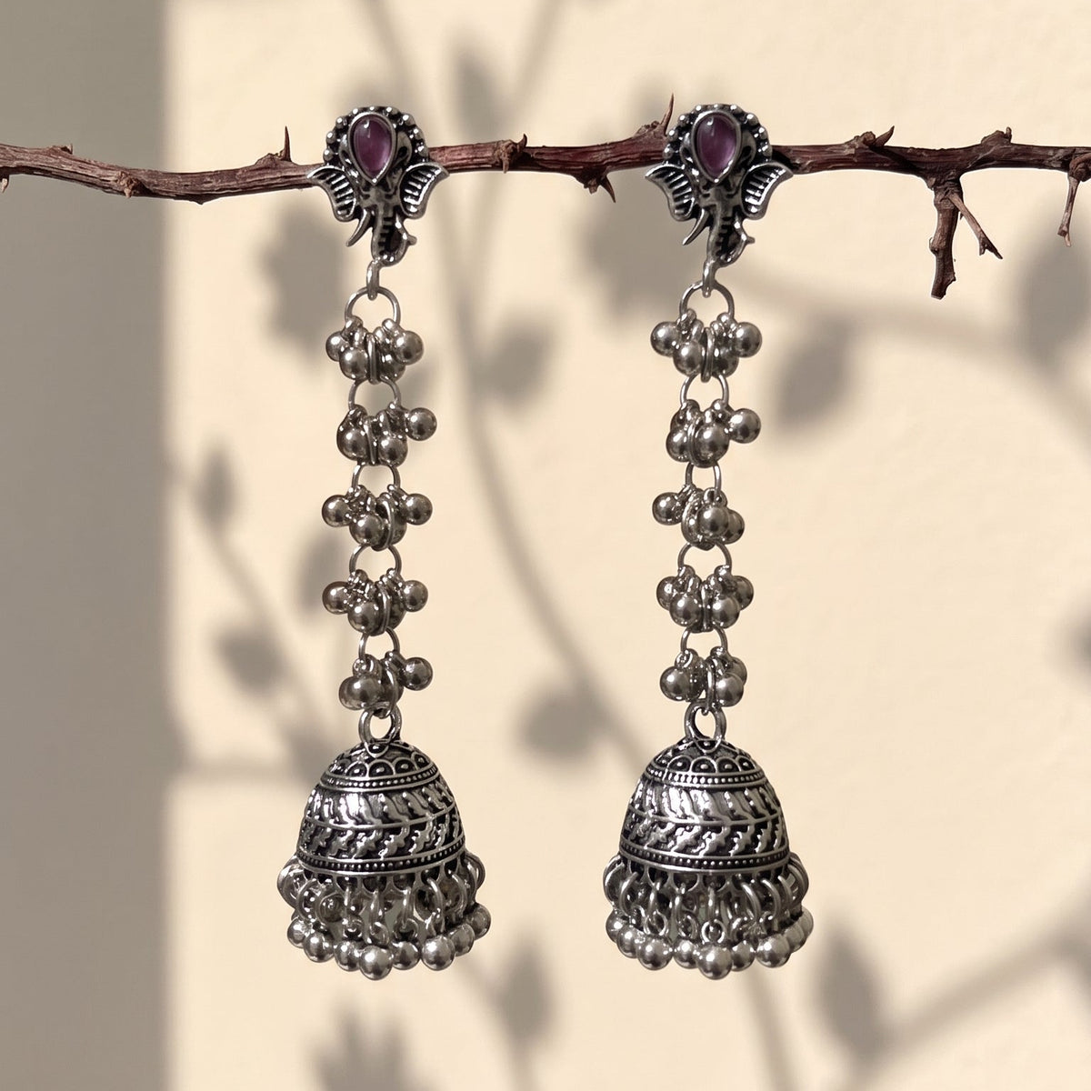 A pair of silver-toned Indian jhumka earrings with small purple stones at the top, hanging from a thin brown branch against a warm beige wall with leaf shadows.
