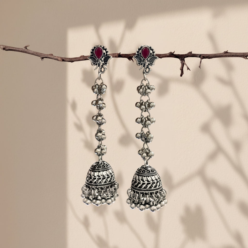 A pair of traditional Indian silver-toned dangle earrings with red oval stones and intricate bell-shaped charms, hanging from a thin branch against a warm beige background with soft leaf shadows.