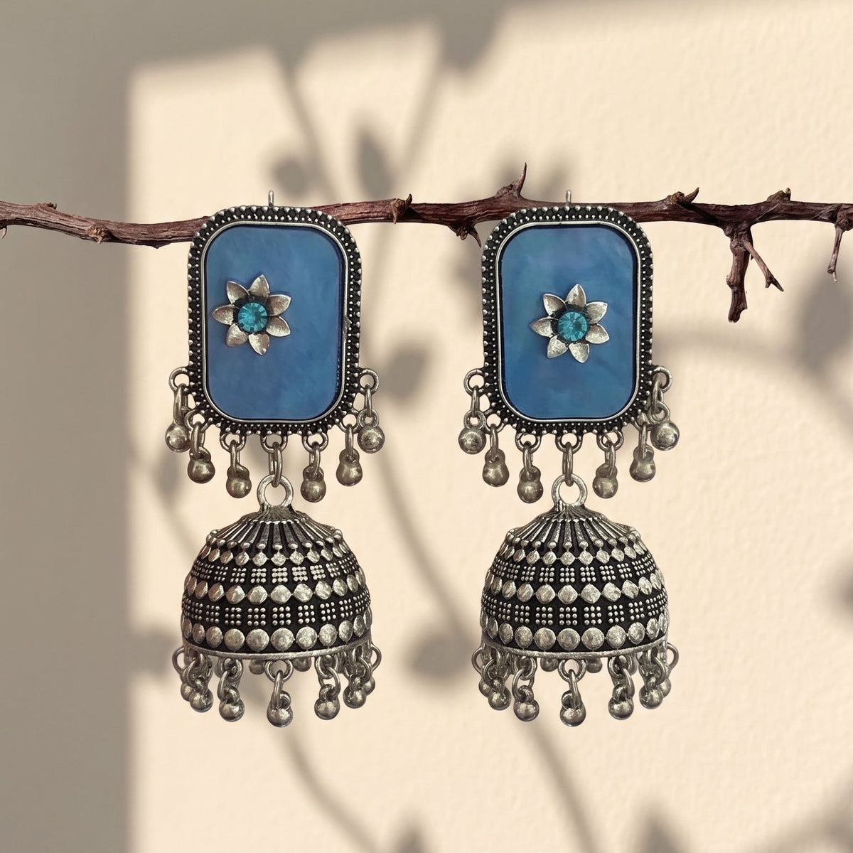 A pair of silver-toned Indian jhumka earrings with rectangular blue stone panels and small turquoise-like blue center stones, hanging from a thin branch against a warm beige wall with leaf shadows.
