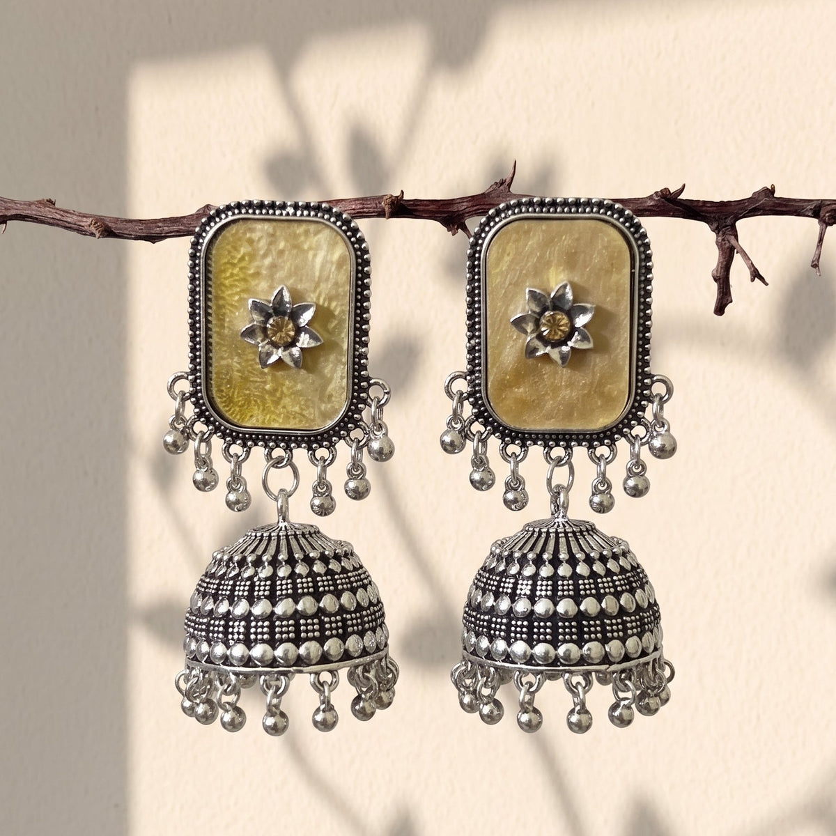 A pair of traditional Indian silver jhumka earrings with rectangular gold-toned mother-of-pearl inlays and dangling silver bells, hanging from a thin branch against a beige wall with leaf shadows.