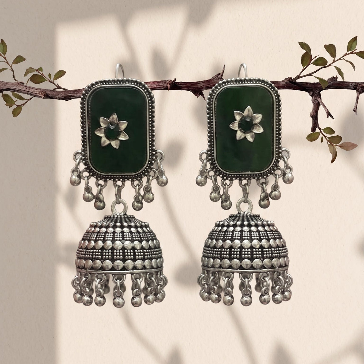 A pair of traditional Indian jhumka earrings hanging from a decorative branch. The earrings feature dark green rectangular inlays with white floral motifs, surrounded by silver-toned borders and dangling silver-toned bell-shaped charms with bead fringe.
