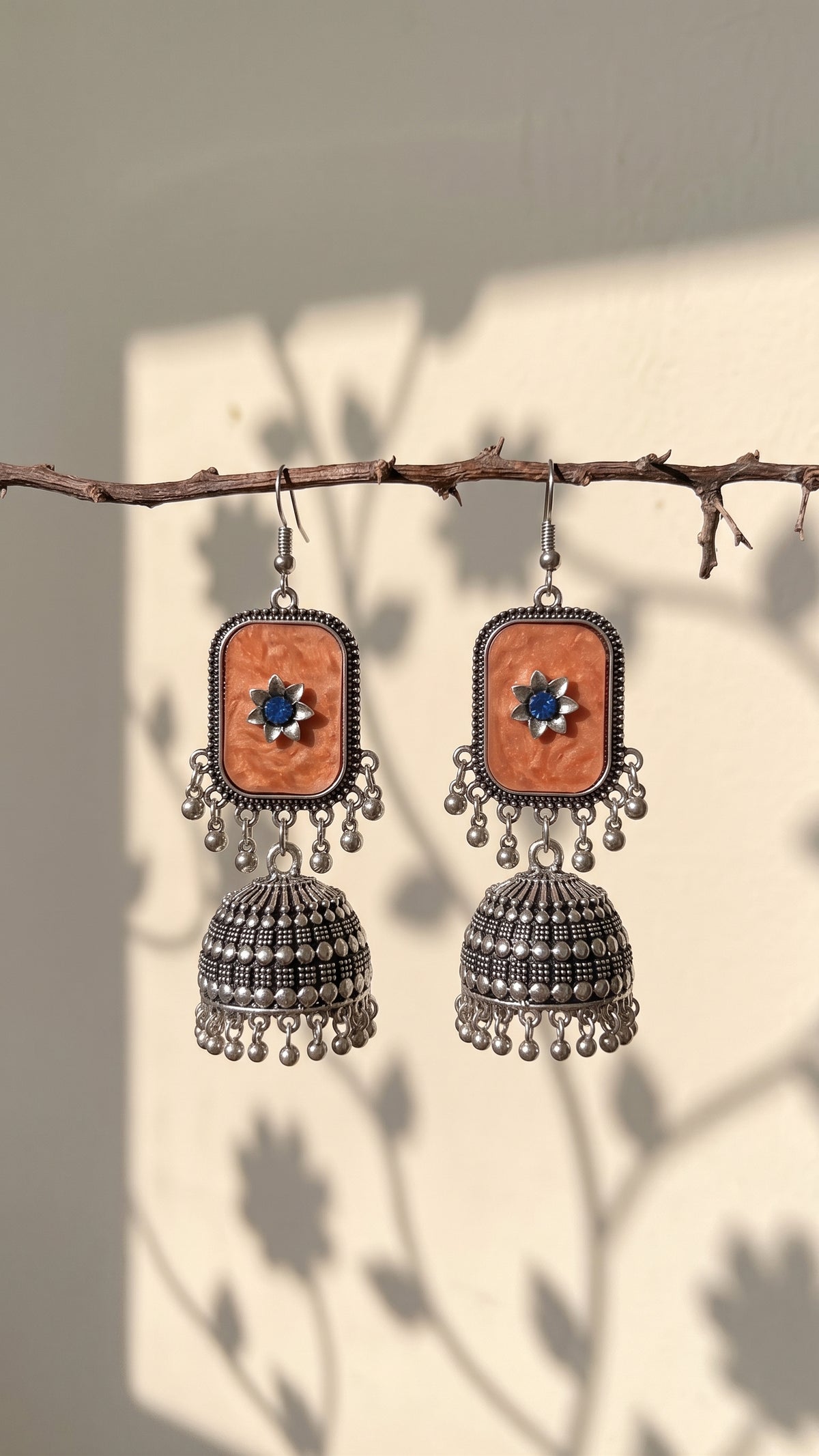 A pair of bohemian-style silver jhumka earrings with rectangular orange stone inlays and blue central stones, hanging from a rustic branch against a wall with leaf shadows.