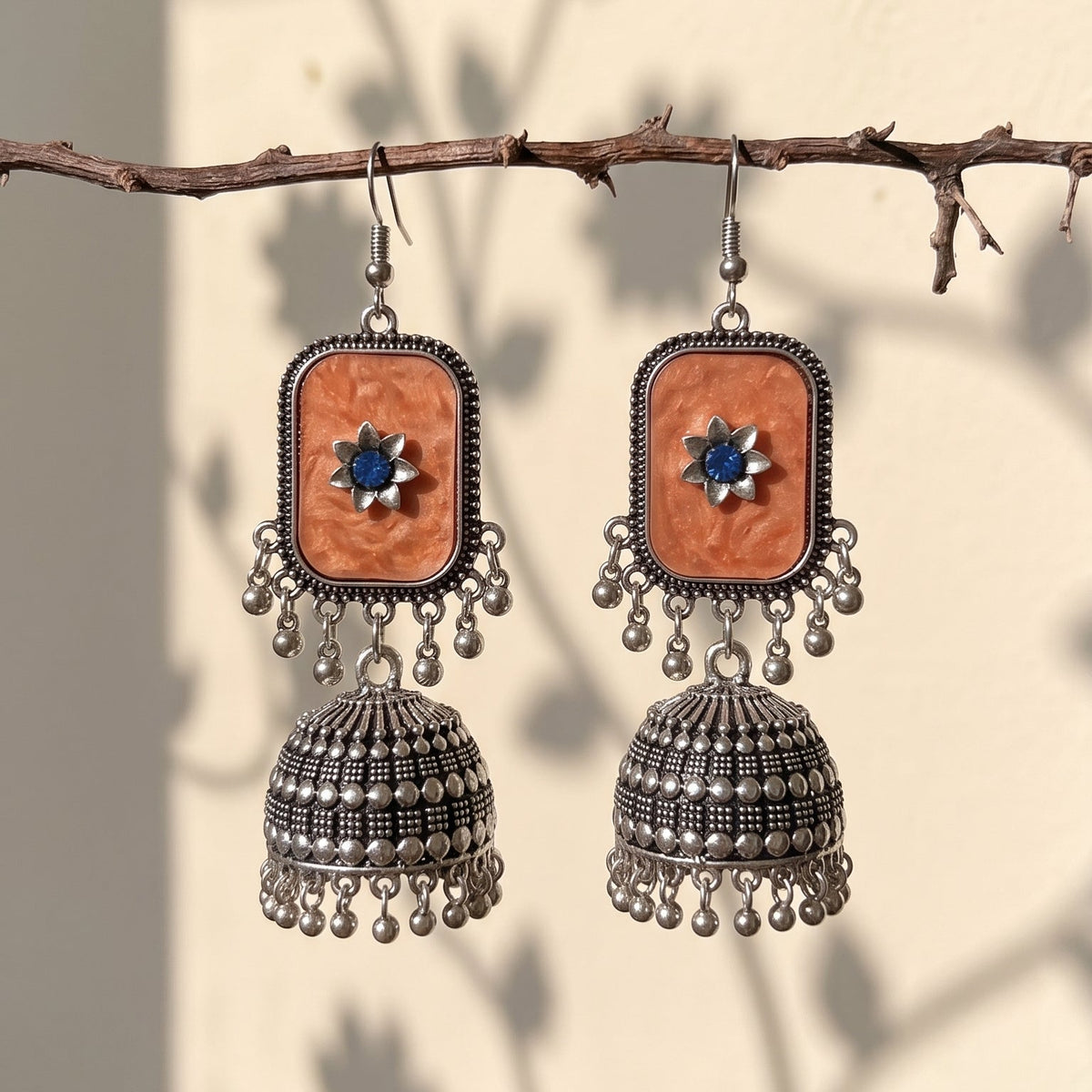 A pair of bohemian-style silver jhumka earrings with rectangular orange stone inlays and blue central stones, hanging from a rustic branch against a wall with leaf shadows.