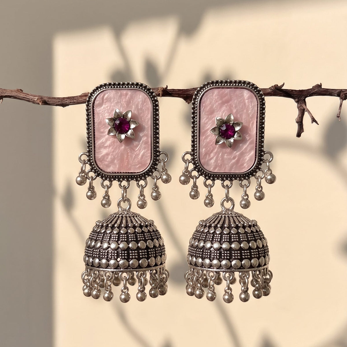 A pair of traditional Indian jhumka earrings with rectangular pink stone studs and silver-toned bell-shaped dangling elements, hanging from a thin branch against a warm beige wall with soft leaf shadows.