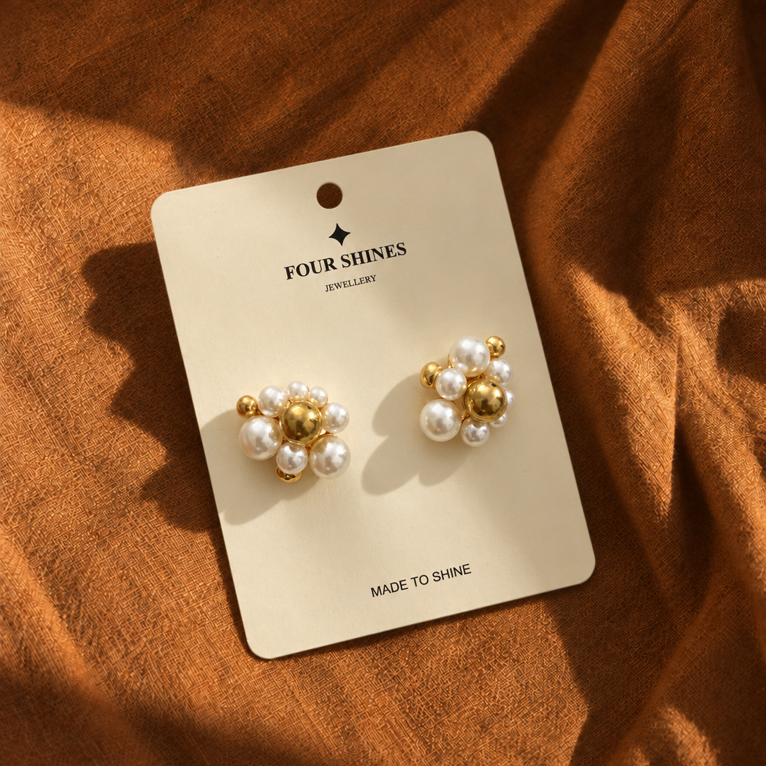 A pair of gold and pearl stud earrings displayed on a cream card labeled FOUR SHINES JEWELLERY, placed on a textured rust-brown fabric background.