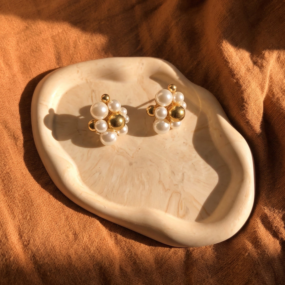 A pair of gold and white pearl stud earrings displayed on a small organic-shaped cream tray, set against a draped brown fabric background with warm sunlight and shadows.