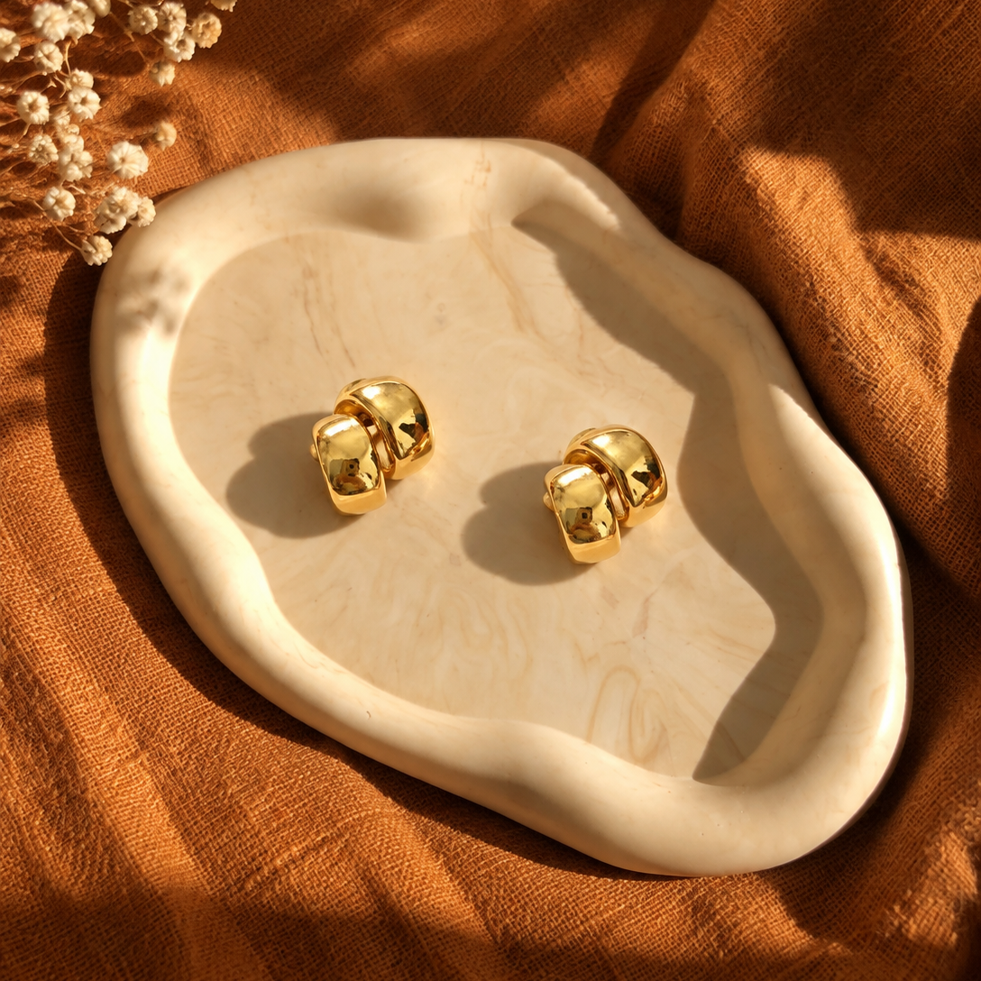 A pair of shiny gold stud earrings displayed on an organic-shaped cream stone tray, set against a warm brown linen fabric background with dried flowers in the corner.