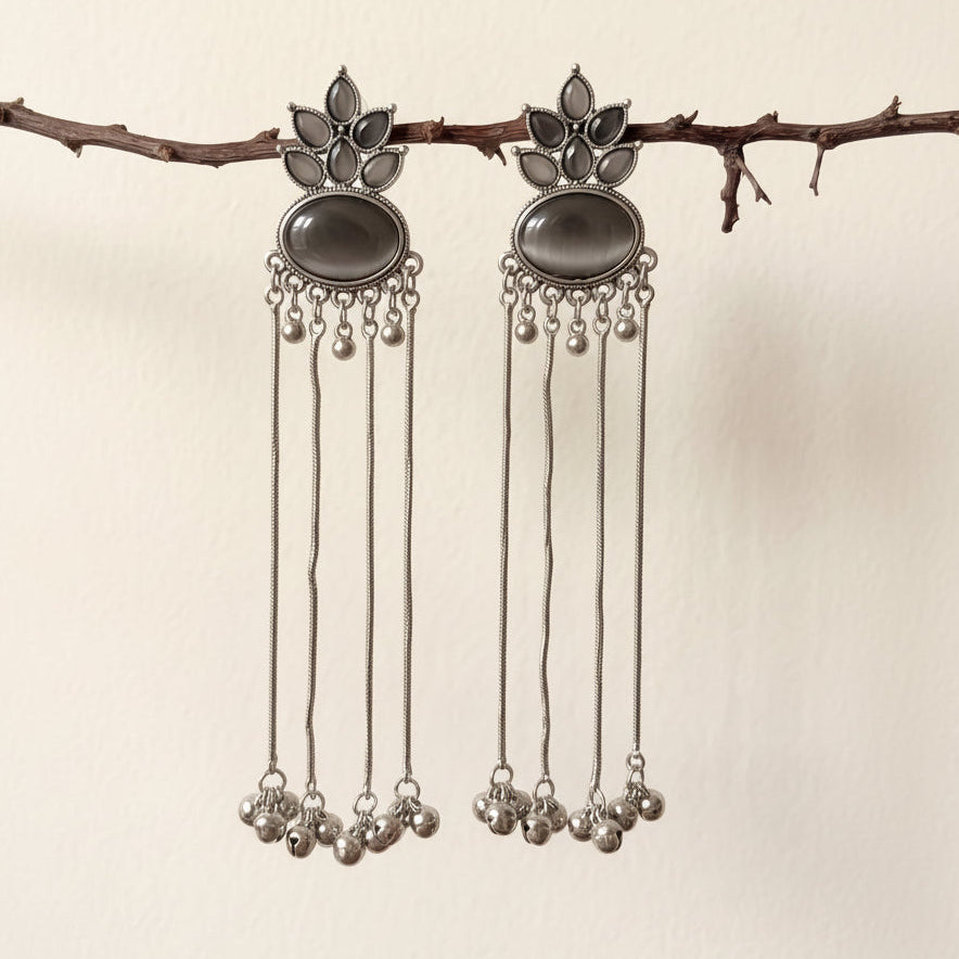 A pair of silver-toned dangle earrings with dark oval stones and dangling chain bells, hanging from a small branch against a plain light beige wall.
