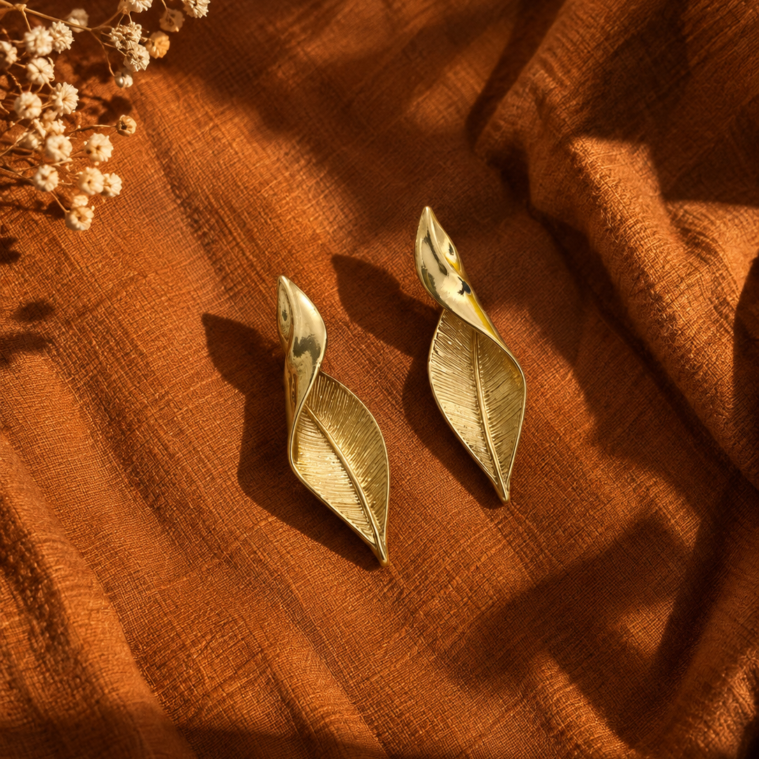 A pair of gold-toned leaf-shaped earrings displayed on draped rust-orange fabric with dried flowers in the corner.