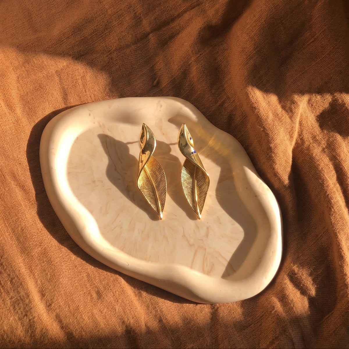 A pair of gold-toned leaf-shaped drop earrings resting on an organic-shaped cream ceramic tray, set against a draped brown fabric background with warm sunlight and shadows.