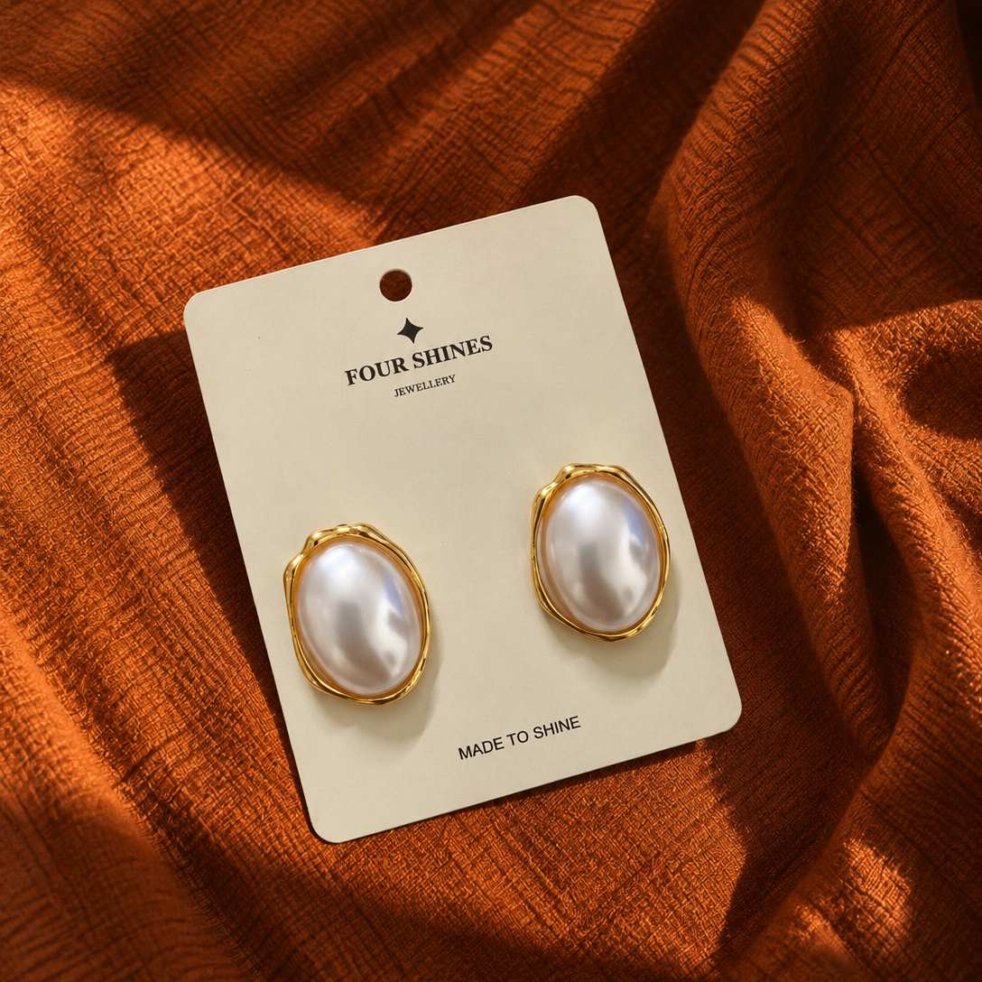 A pair of oval pearl stud earrings with gold bezel settings, mounted on a cream-colored card labeled 'FOUR SHINES JEWELRY', placed on a draped rust-orange fabric background.