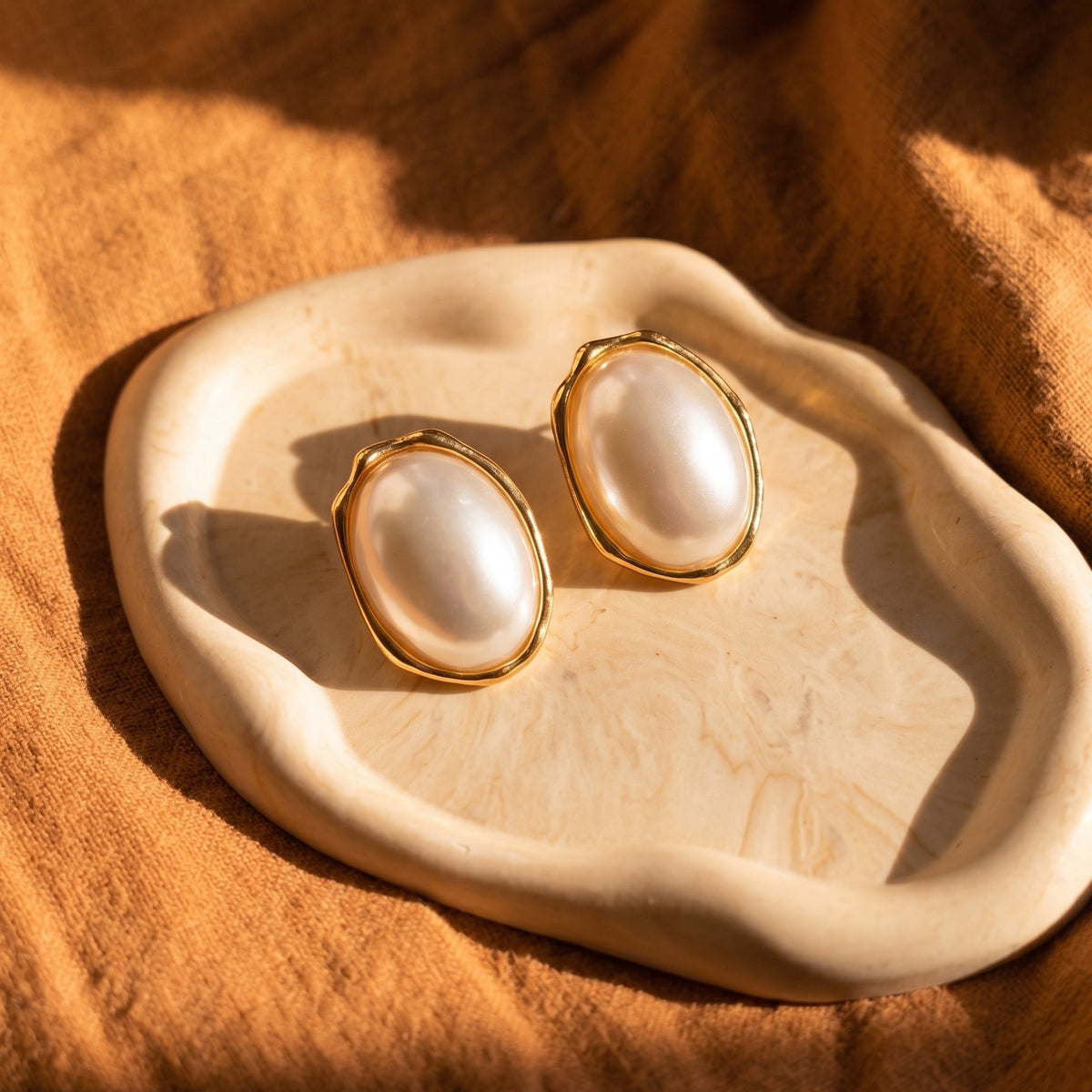 A pair of gold-tone stud earrings with large oval white pearl-like centers, displayed on a cream ceramic dish over a warm brown fabric background under dramatic sunlight.