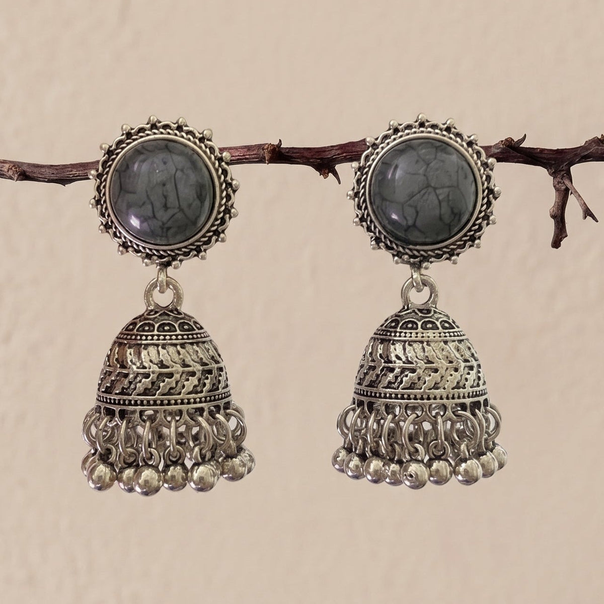 A pair of traditional Indian silver jhumka earrings with oval dark gray stones, hanging from a thin brown branch against a textured beige wall.