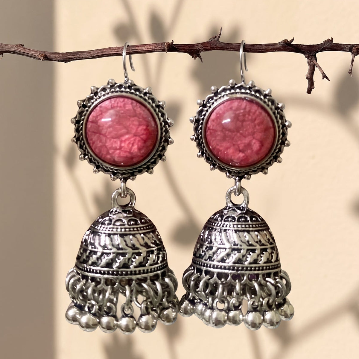 A pair of silver-toned Indian jhumka earrings with round pink cabochon stones, hanging from a thin brown branch against a beige wall with leaf shadows.