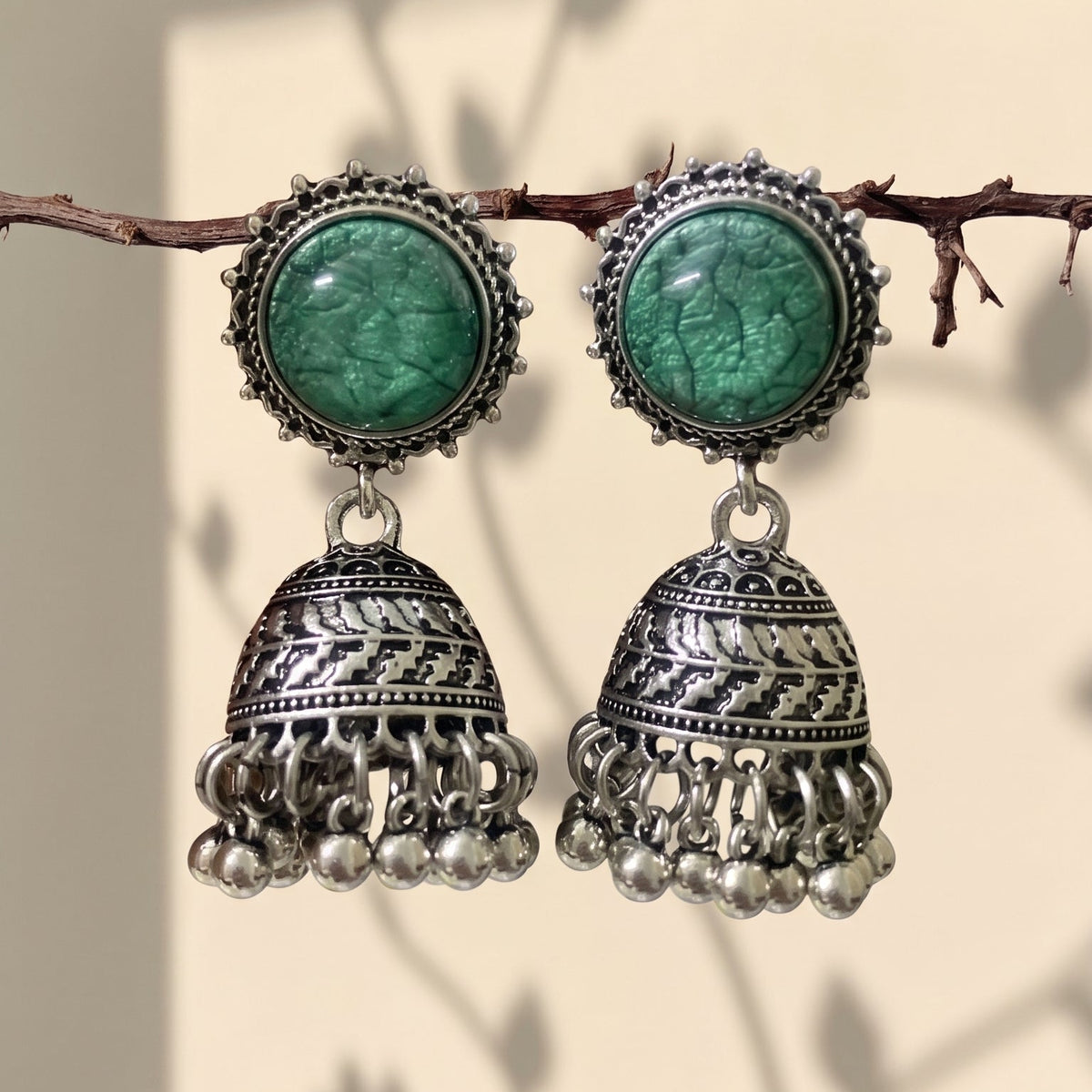 A pair of traditional Indian silver jhumka earrings with green stone studs, hanging from a thin branch against a warm beige background with soft leaf shadows.