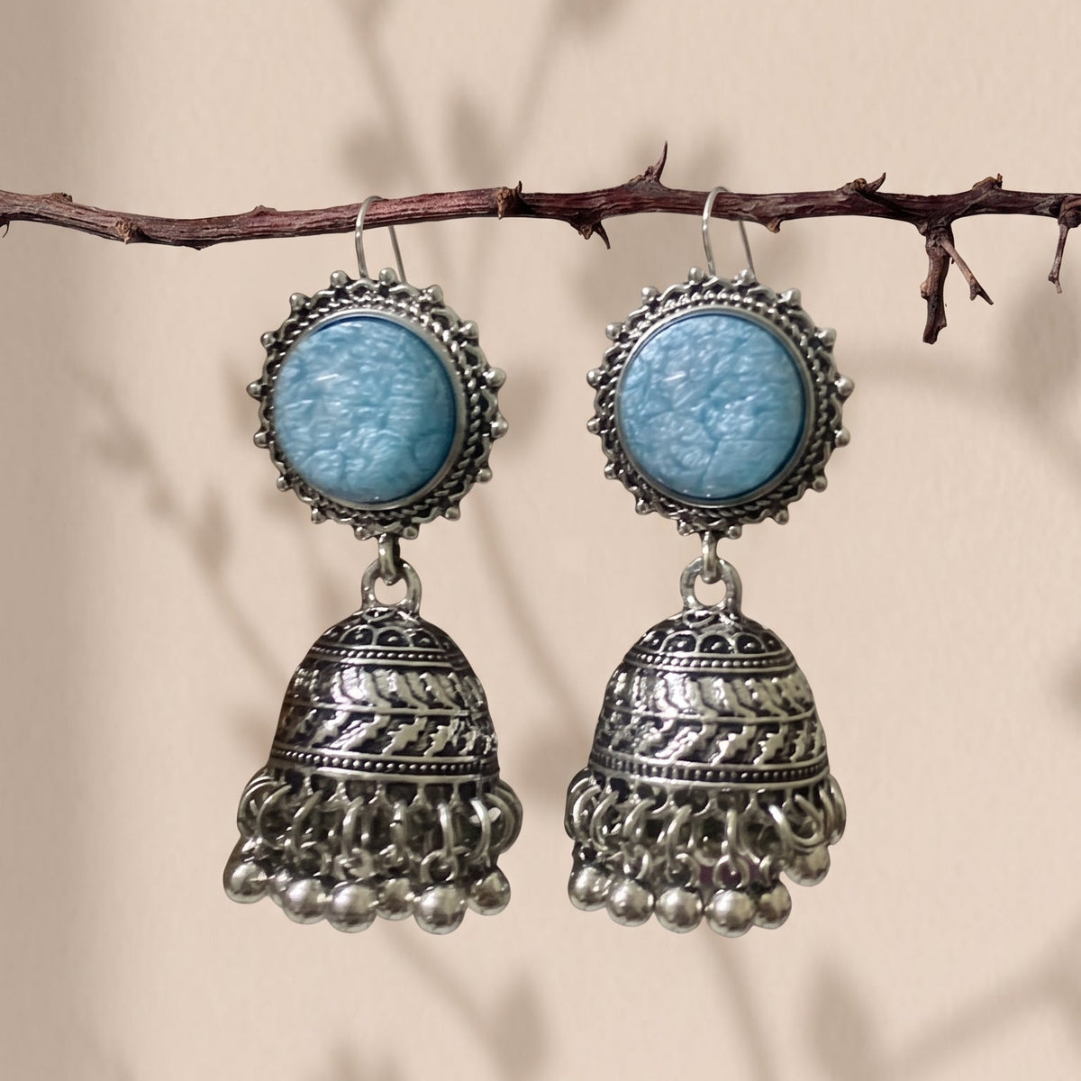 A pair of bohemian silver jhumka earrings with light blue cabochon stones, hanging from a thin brown branch against a beige background with soft leaf shadows.