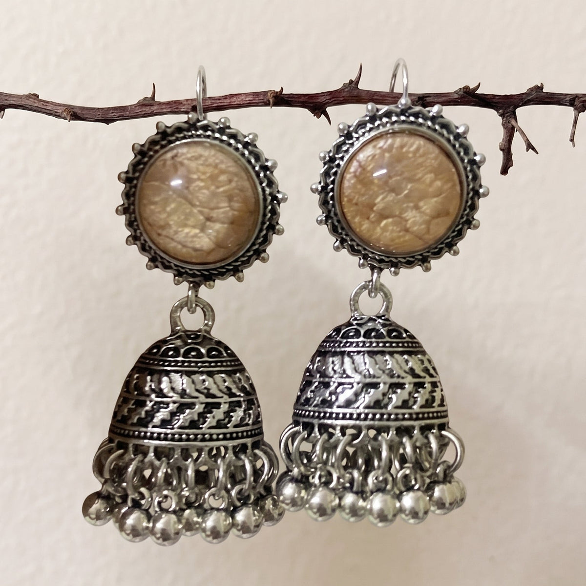 A pair of traditional Indian silver-toned jhumka earrings with oval beige stone inlays, hanging from a small branch against a plain beige wall.