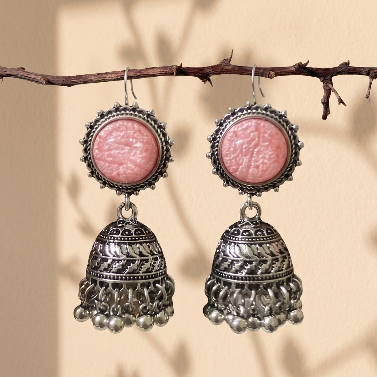A pair of silver-toned jhumka earrings with round pink cabochon stones, hanging from a thin branch against a warm beige background with soft leaf shadows.