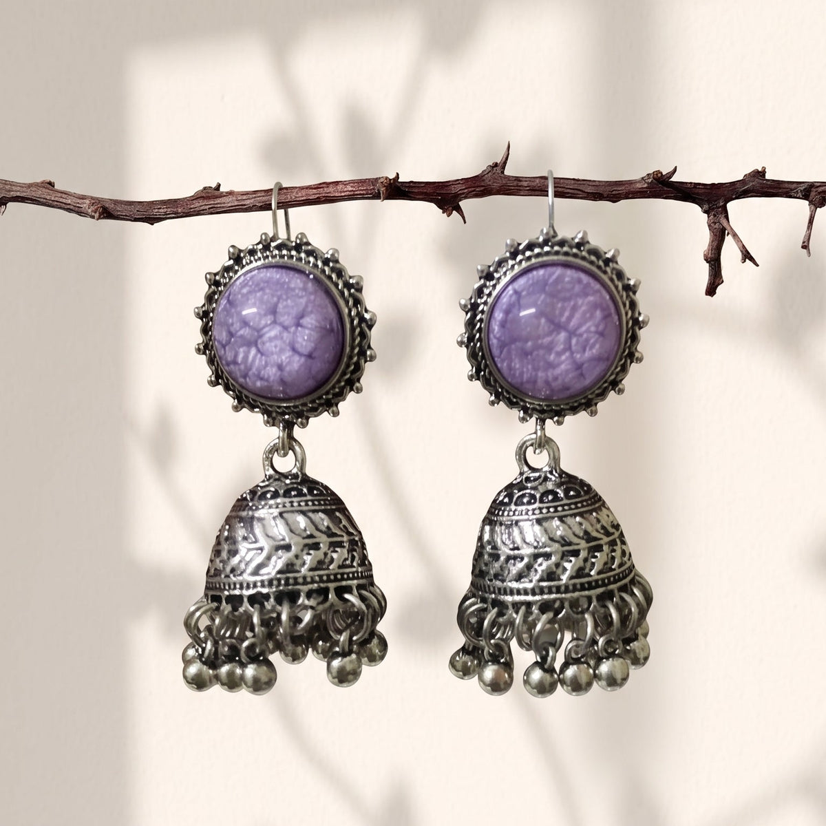 A pair of silver-toned jhumka earrings with oval purple stone tops, hanging from a thin branch against a warm beige background with soft leaf shadows.