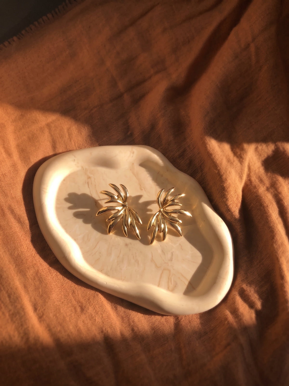 A pair of gold-toned, palm-leaf-shaped stud earrings displayed on an organic-shaped cream ceramic tray, set against a draped rust-orange linen fabric background.