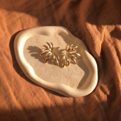 A pair of gold-toned, palm-leaf-shaped stud earrings displayed on an organic-shaped cream ceramic tray, set against a draped rust-orange linen fabric background.