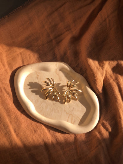 A pair of gold-toned, palm-leaf-shaped stud earrings displayed on an organic-shaped cream ceramic tray, set against a draped rust-orange linen fabric background.