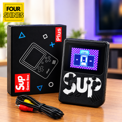 A retro-style handheld game console with a small screen and white 'sup' branding, shown next to its black retail box on a wooden table. The box has blue geometric shapes and red 'Plus' and 'Sup Game Box' labels.