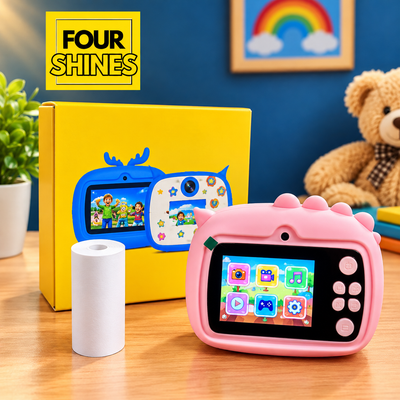 A pink children's digital camera with a colorful screen and dinosaur-themed handle, shown in front of its yellow box and a white roll of film. The background features a blue wall, a framed rainbow, and a teddy bear, with a 'FOUR SHINES' logo in the top left corner.