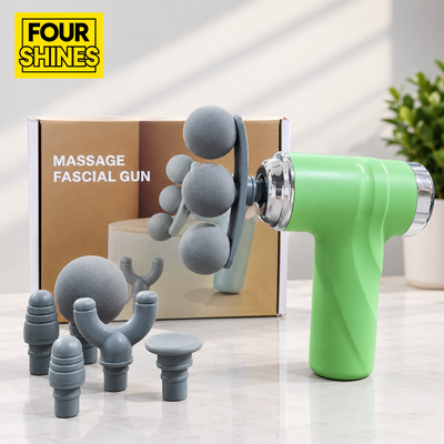 A green handheld massage facial gun with multiple gray silicone massage heads and an included cardboard box, photographed on a light marble surface with a plant in the background. The yellow FOUR SHINES logo appears in the top-left corner.