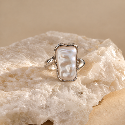 A silver ring with a large, irregularly shaped white mother-of-pearl cabochon, displayed on a textured white crystal cluster against a warm beige background.