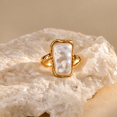 A gold ring with a large, irregularly shaped white pearl bezel-set on a textured white stone, photographed against a warm beige background.